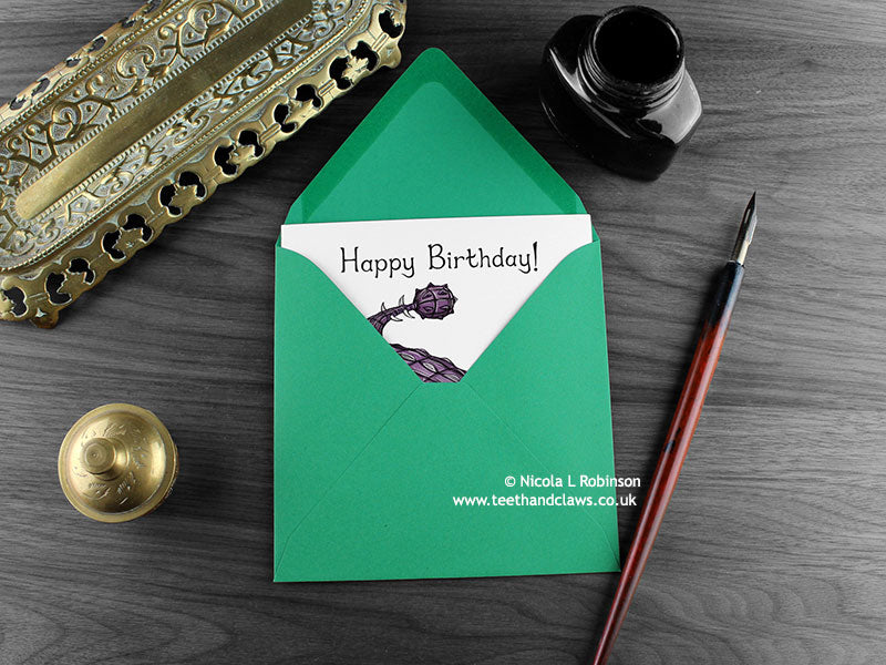Dinosaur birthday card, shown lying flat, with my original pen and ink illustration of an Ankylosaurus, inside an open green envelope. With vintage Victorian pen and writing desk accessories. Design and illustration © Nicola L Robinson | Teeth and Claws