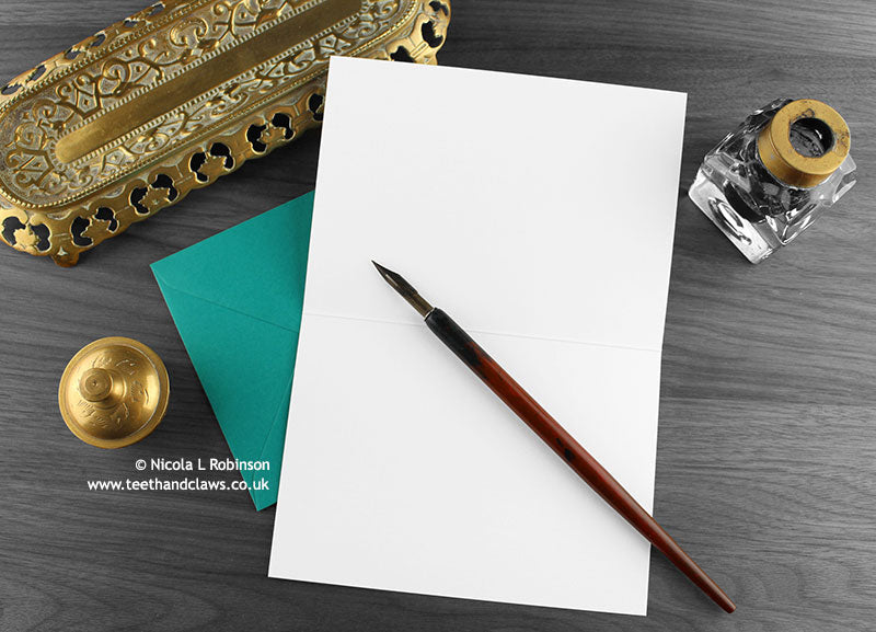 British shorthair cat greeting card, shown open and flat with blank inside, on a teal envelope, alongside vintage Victorian pen and writing desk accessories. Design and illustration © Nicola L Robinson | Teeth and Claws