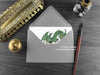 Green Dragon New Baby Card, shown lying flat, with my original pen and ink illustration of a baby green dragon, inside an open grey envelope. With vintage Victorian pen and writing desk accessories. Design and illustration © Nicola L Robinson | Teeth and Claws