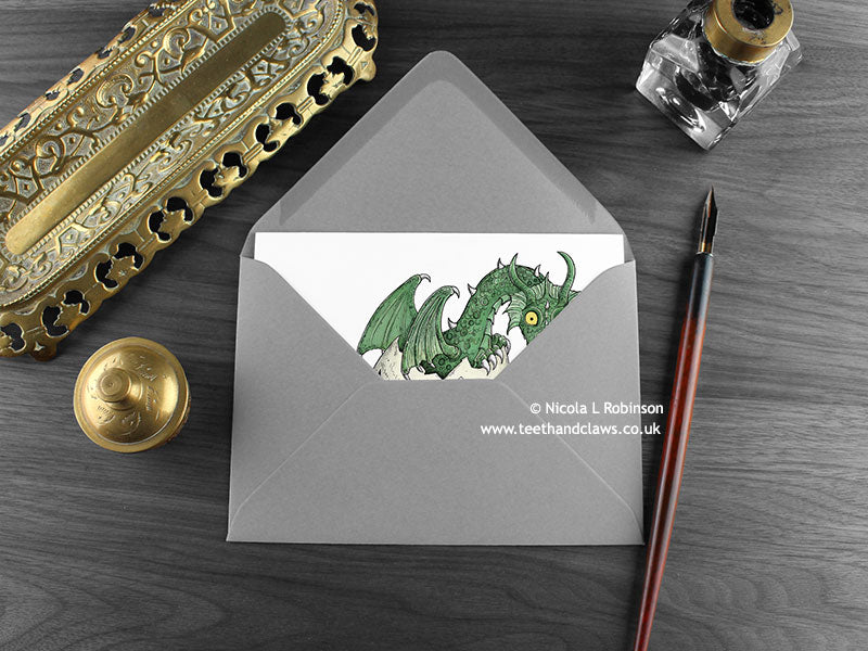 Green Dragon New Baby Card, shown lying flat, with my original pen and ink illustration of a baby green dragon, inside an open grey envelope. With vintage Victorian pen and writing desk accessories. Design and illustration © Nicola L Robinson | Teeth and Claws