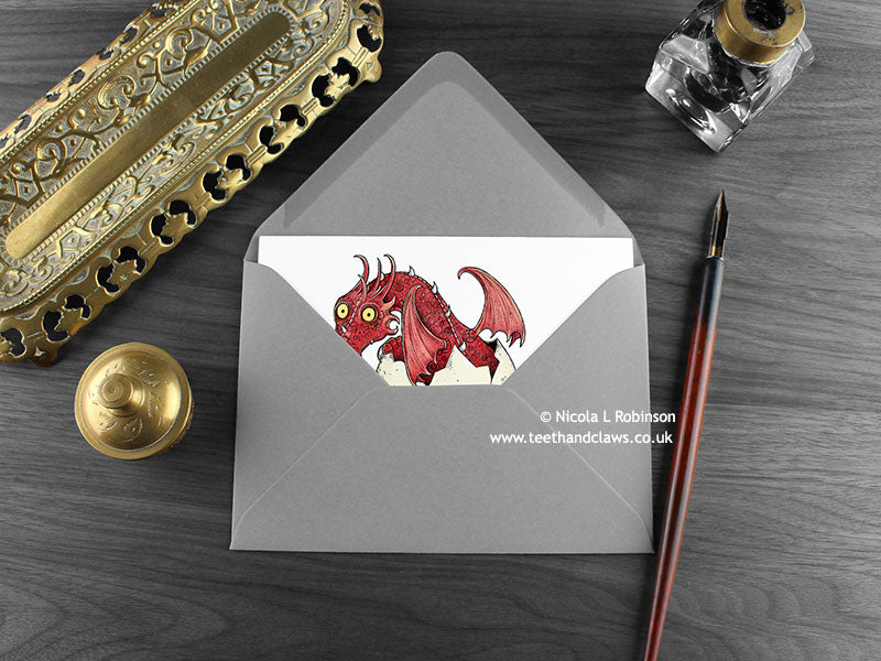 Red Dragon New Baby Card, shown lying flat, with my original pen and ink illustration of a baby red dragon, inside an open grey envelope. With vintage Victorian pen and writing desk accessories. Design and illustration © Nicola L Robinson | Teeth and Claws