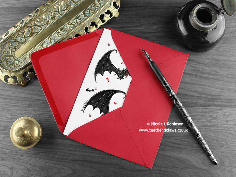 Gothic bats love card, shown lying flat, with my original pen and ink illustration of Gothic bats, inside an open red envelope. With vintage Victorian pen and writing desk accessories. Design and illustration © Nicola L Robinson | Teeth and Claws