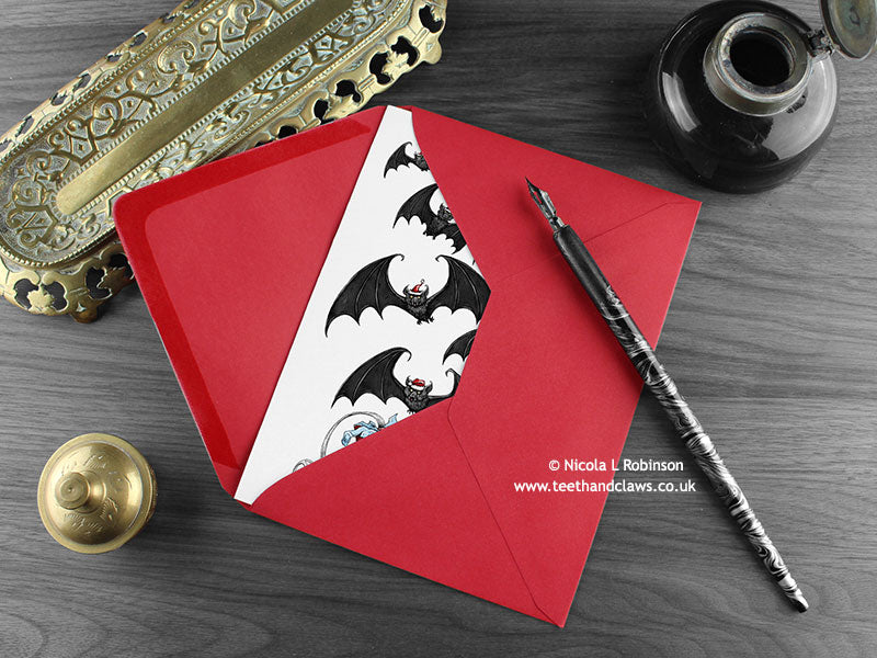 Gothic Bats Christmas Card, shown lying flat, with my original pen and ink illustration of festive bats, inside an open red envelope. With vintage Victorian pen and writing desk accessories. Design and illustration © Nicola L Robinson | Teeth and Claws