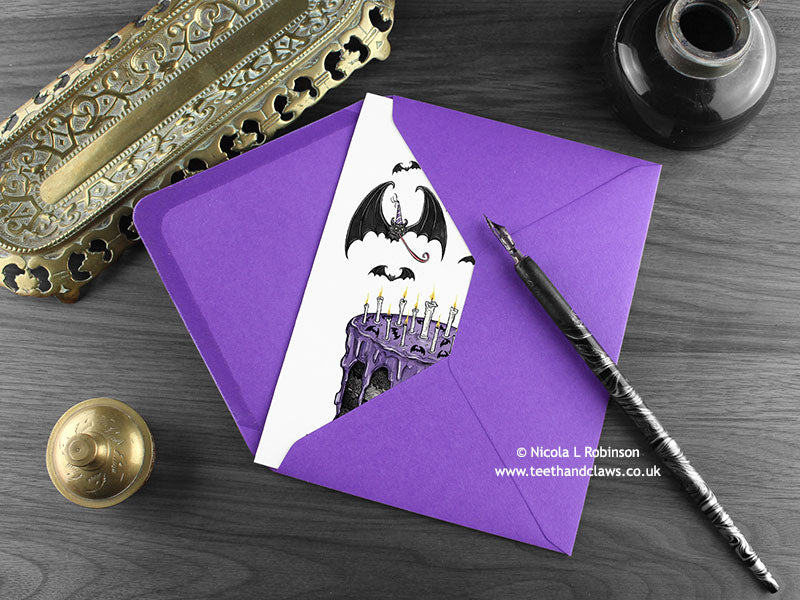 Gothic Bat Birthday cake card, shown lying flat, with my original pen and ink illustration of bats and birthday cake, inside an open purple envelope. With vintage Victorian pen and writing desk accessories. Design and illustration © Nicola L Robinson | Teeth and Claws