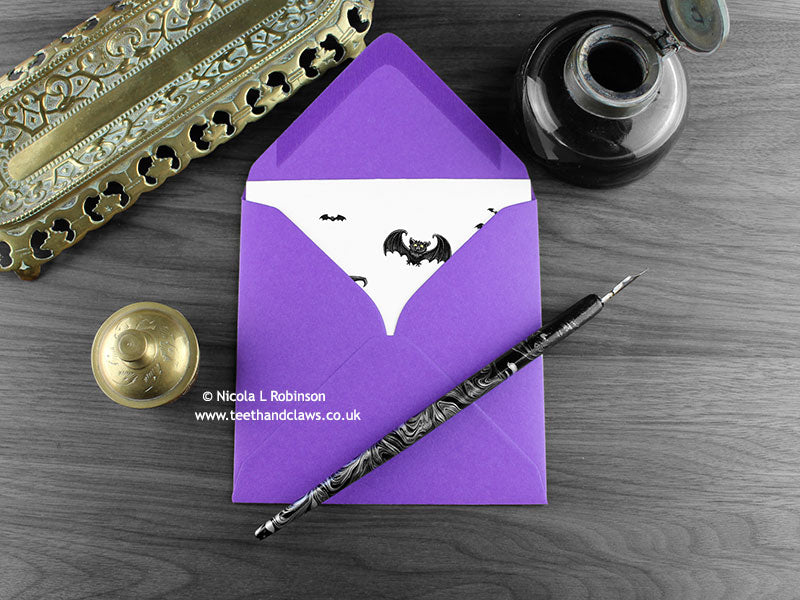 Gothic Bat Mother’s Day, New Baby Card, shown lying flat, with my original pen and ink illustration of flying bats, inside an open purple envelope. With vintage Victorian pen and writing desk accessories. Design and illustration © Nicola L Robinson | Teeth and Claws