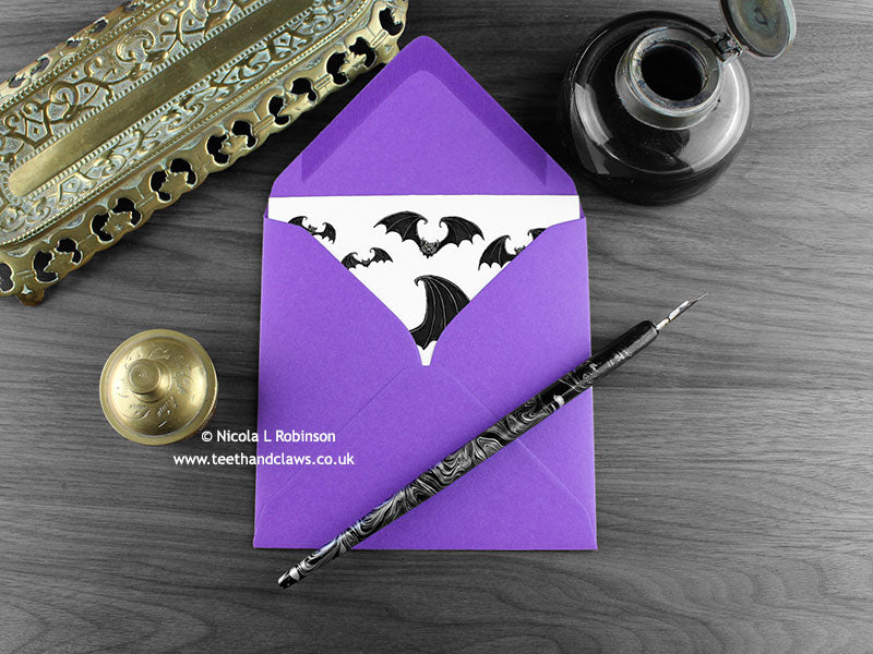 Gothic bats greeting card, shown lying flat, with my original pen and ink illustration of flying bats, inside an open purple envelope. With vintage Victorian pen and writing desk accessories. Design and illustration © Nicola L Robinson | Teeth and Claws