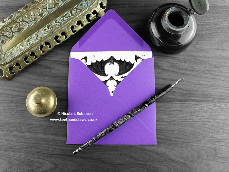 Gothic bats greeting card, shown lying flat, with my original pen and ink illustration of flying bats, inside an open purple envelope. With vintage Victorian pen and writing desk accessories. Design and illustration © Nicola L Robinson | Teeth and Claws