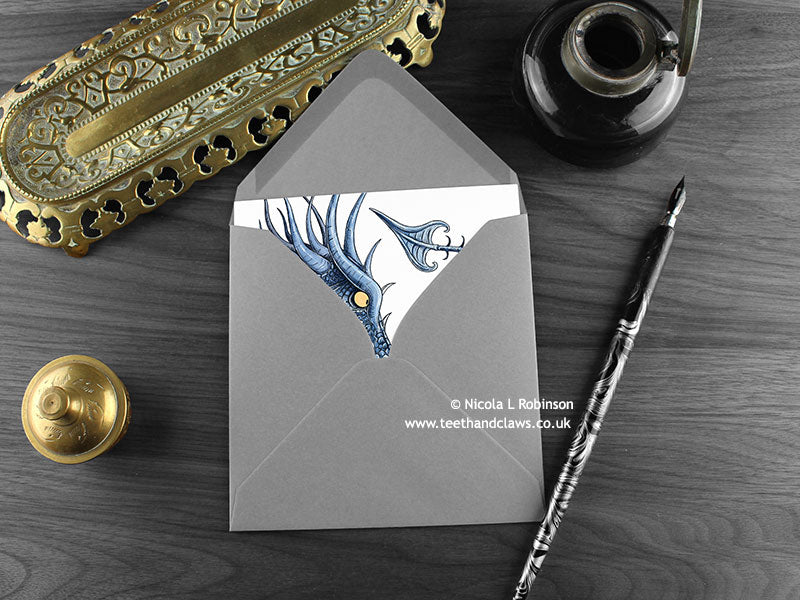 Big Blue Dragon and Little Green Dragon Greeting Card, shown lying flat, with my original pen and ink illustration of a big blue dragon, inside an open grey envelope. With vintage Victorian pen and writing desk accessories. Design and illustration © Nicola L Robinson | Teeth and Claws