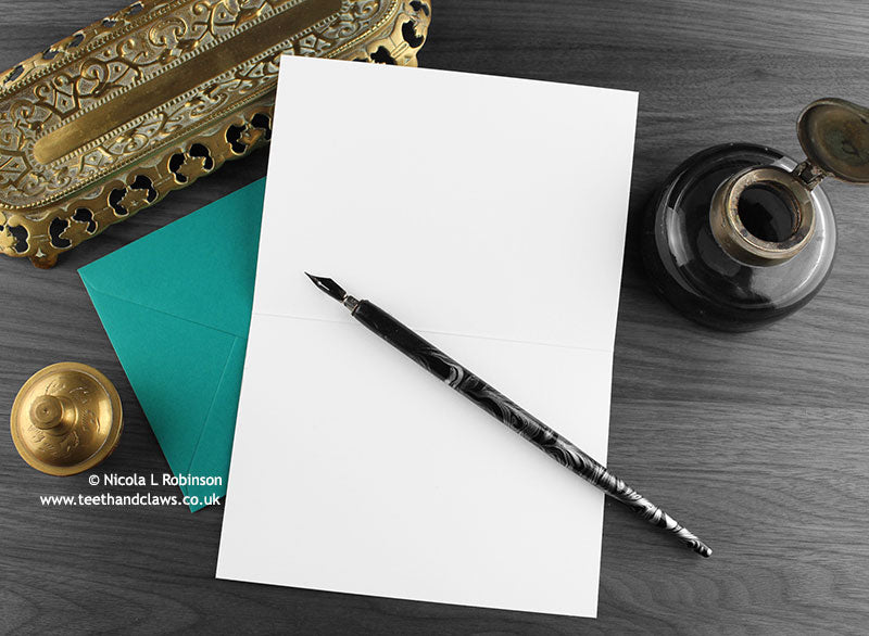 Black cat greeting card, shown open and flat with blank inside, on a teal envelope, alongside vintage Victorian pen and writing desk accessories. Design and illustration © Nicola L Robinson | Teeth and Claws
