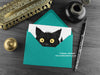 Black cat greeting card, shown lying flat, with my original pen and ink illustration of a black cat, inside an open teal envelope. With vintage Victorian pen and writing desk accessories. Design and illustration © Nicola L Robinson | Teeth and Claws