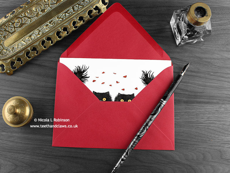 Black Persian cats love card, shown lying flat, with my original pen and ink illustration of black Persian cats, inside an open red envelope. With vintage Victorian pen and writing desk accessories. Design and illustration © Nicola L Robinson | Teeth and Claws
