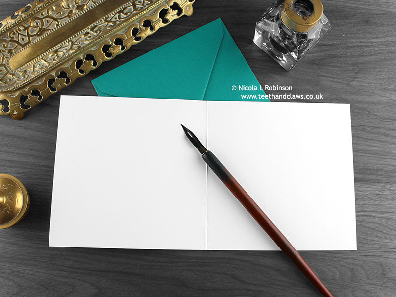 Black and white cat greeting card, shown open and flat with blank inside, on a teal envelope, alongside vintage Victorian pen and writing desk accessories. Design and illustration © Nicola L Robinson | Teeth and Claws