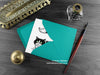 Black and white cat greeting card, shown lying flat, with my original pen and ink illustration of a tuxedo cat, inside an open teal envelope. With vintage Victorian pen and writing desk accessories. Design and illustration © Nicola L Robinson | Teeth and Claws