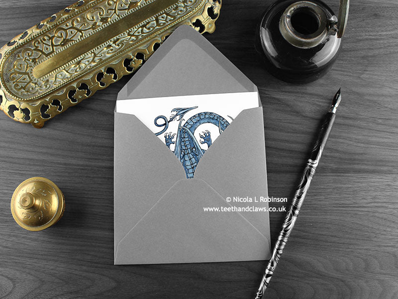 Blue dragon greeting card, shown lying flat, with my original pen and ink illustration of a blue dragon, inside an open grey envelope. With vintage Victorian pen and writing desk accessories. Design and illustration © Nicola L Robinson | Teeth and Claws