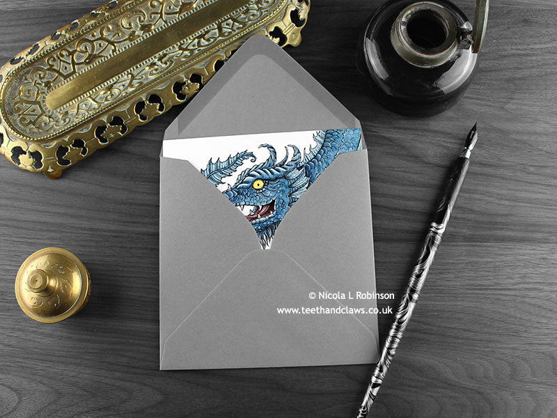 Blue Serpent Dragon Greeting Card, shown lying flat, with my original pen and ink illustration of a blue dragon, inside an open grey envelope. With vintage Victorian pen and writing desk accessories. Design and illustration © Nicola L Robinson | Teeth and Claws