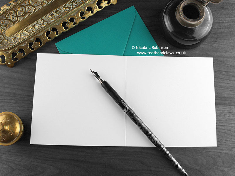 British Shorthair cat greeting card, shown open and flat with blank inside, on a teal envelope, alongside vintage Victorian pen and writing desk accessories. Design and illustration © Nicola L Robinson | Teeth and Claws