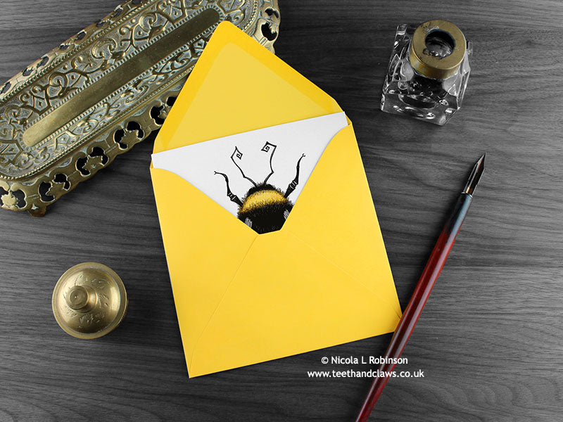 Bee greeting card, shown lying flat, with my original pen and ink illustration of a bumble bee, inside an open yellow envelope. With vintage Victorian pen and writing desk accessories. Design and illustration © Nicola L Robinson | Teeth and Claws
