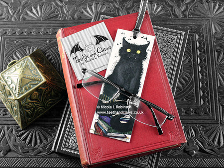Raven bookmark featuring my original pen and ink illustration, front view of the bookmark resting on books with one of my branded cards. Made in the UK. Design and illustration © Nicola L Robinson | Teeth and Claws.