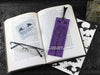 Black cat bookmark featuring my original pen and ink illustration, back view of the bookmark resting on an open book with reading glasses and one of my branded cards. Made in the UK. Design and illustration © Nicola L Robinson | Teeth and Claws.