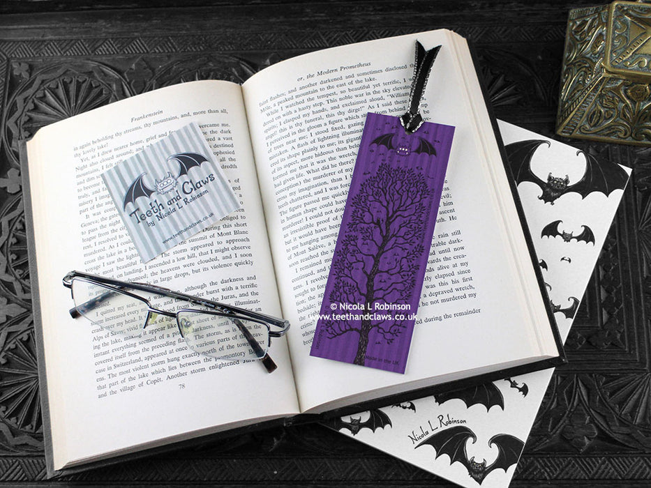Black cat bookmark featuring my original pen and ink illustration, back view of the bookmark resting on an open book with reading glasses and one of my branded cards. Made in the UK. Design and illustration © Nicola L Robinson | Teeth and Claws.