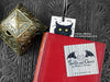 Black cat bookmark featuring my original pen and ink illustration, front view with the top showing from inside a closed book with one of my branded cards. Made in the UK. Design and illustration © Nicola L Robinson | Teeth and Claws.