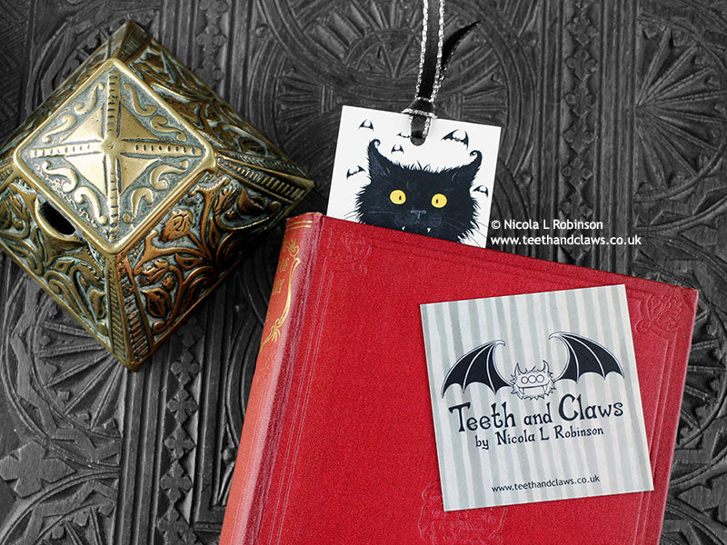 Black cat bookmark featuring my original pen and ink illustration, front view with the top showing from inside a closed book with one of my branded cards. Made in the UK. Design and illustration © Nicola L Robinson | Teeth and Claws.