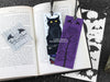 Black Cat bookmark featuring my original pen and ink illustrations, front and back view of the bookmark resting on an open book with one of my branded cards. Made in the UK. Design and illustration © Nicola L Robinson | Teeth and Claws.
