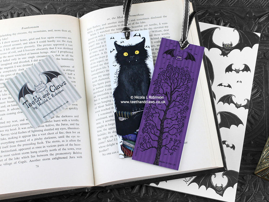 Black Cat bookmark featuring my original pen and ink illustrations, front and back view of the bookmark resting on an open book with one of my branded cards. Made in the UK. Design and illustration © Nicola L Robinson | Teeth and Claws.