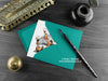 Calico cat greeting card, shown lying flat, with my original pen and ink illustration of a calico cat, inside an open teal envelope. With vintage Victorian pen and writing desk accessories. Design and illustration © Nicola L Robinson | Teeth and Claws