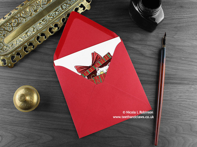 Cats Christmas card, shown lying flat, with my original pen and ink illustration of two  festive cats, inside an open red envelope. With vintage Victorian pen and writing desk accessories. Design and illustration © Nicola L Robinson | Teeth and Claws