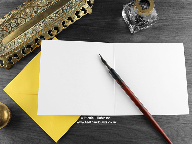 Cat and chick greeting card, shown open and flat with blank inside, on a yellow envelope, alongside vintage Victorian pen and writing desk accessories. Design and illustration © Nicola L Robinson | Teeth and Claws