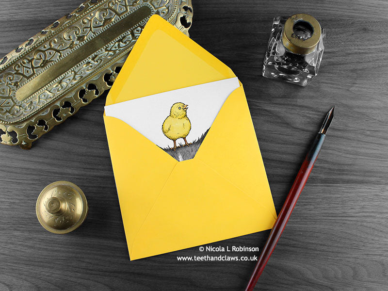 Cat and chick greeting card, shown lying flat, with my original pen and ink illustration of a cat and chick, inside an open yellow envelope. With vintage Victorian pen and writing desk accessories. Design and illustration © Nicola L Robinson | Teeth and Claws