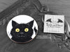 Black Cat Coaster featuring my original pen and ink illustration of a black cat face, front view alongside one of my branded cards. Made in the UK. Design and illustration © Nicola L Robinson | Teeth and Claws.