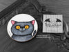 British Shorthair Cat Coaster featuring my original pen and ink illustration of a grey British Shorthair cat face, front view alongside one of my branded cards. Made in the UK. Design and illustration © Nicola L Robinson | Teeth and Claws.