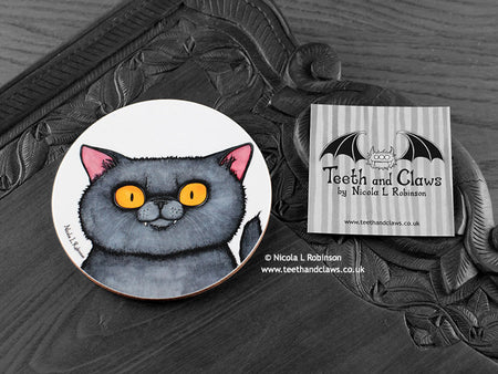 British Shorthair Cat Coaster featuring my original pen and ink illustration of a grey British Shorthair cat face, front view alongside one of my branded cards. Made in the UK. Design and illustration © Nicola L Robinson | Teeth and Claws.