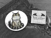Maine Coon Cat Coaster featuring my original pen and ink illustration of a Maine Coon cat, front view alongside one of my branded cards. Made in the UK. Design and illustration © Nicola L Robinson | Teeth and Claws.