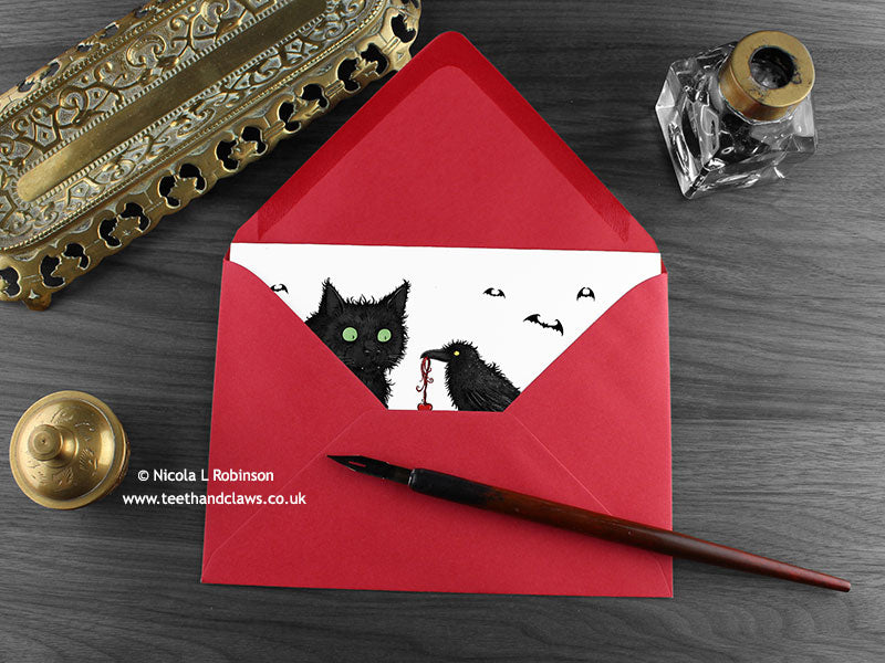 Black cat and crow love card, shown lying flat, with my original pen and ink illustration of a black cat and crow, inside an open red envelope. With vintage Victorian pen and writing desk accessories. Design and illustration © Nicola L Robinson | Teeth and Claws