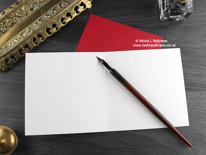 Cat and Robin Christmas card, shown open and flat with blank inside, on a red envelope, alongside vintage Victorian pen and writing desk accessories. Design and illustration © Nicola L Robinson | Teeth and Claws