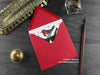 Cat and Robin Christmas card, shown lying flat, with my original pen and ink illustration of a festive cat, inside an open red envelope. With vintage Victorian pen and writing desk accessories. Design and illustration © Nicola L Robinson | Teeth and Claws