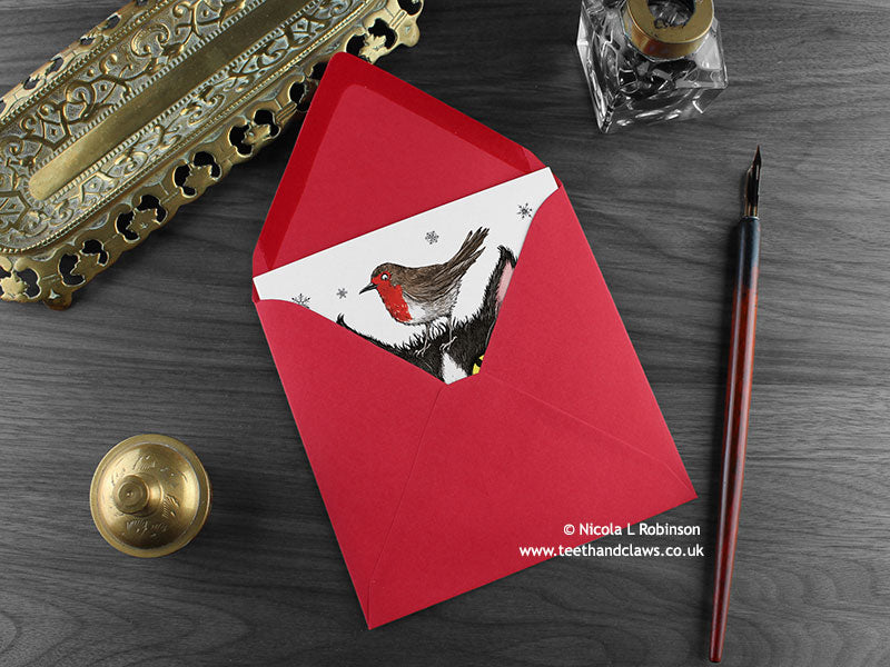 Cat and Robin Christmas card, shown lying flat, with my original pen and ink illustration of a festive cat, inside an open red envelope. With vintage Victorian pen and writing desk accessories. Design and illustration © Nicola L Robinson | Teeth and Claws