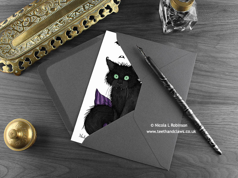 Black cat greeting card, shown lying flat, with my original pen and ink illustration of a black cat, inside an open dark grey envelope. With vintage Victorian pen and writing desk accessories. Design and illustration © Nicola L Robinson | Teeth and Claws