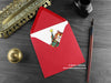 Ginger Cat Christmas card, shown lying flat, with my original pen and ink illustration of a festive cat, inside an open red envelope. With vintage Victorian pen and writing desk accessories. Design and illustration © Nicola L Robinson | Teeth and Claws