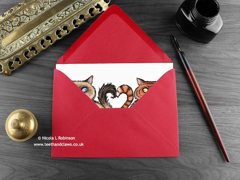 Cat love card, shown lying flat, with my original pen and ink illustration of two cats, inside an open red envelope. With vintage Victorian pen and writing desk accessories. Design and illustration © Nicola L Robinson | Teeth and Claws