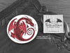 Red Dragon Coaster featuring my original pen and ink illustration of a red dragon, front view alongside one of my branded cards. Made in the UK. Design and illustration © Nicola L Robinson | Teeth and Claws.