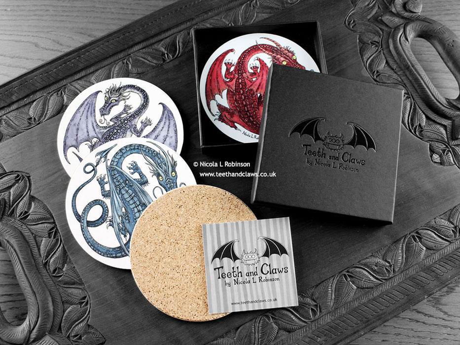 Set of four of my dragon coasters featuring my original pen and ink dragon illustrations, front view alongside a black gift box and card, with one coaster turned over to show the cork backing. Made in the UK. Design and illustration © Nicola L Robinson | Teeth and Claws.