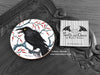 Crow Coaster featuring my original pen and ink illustration of a crow, front view alongside one of my branded cards. Made in the UK. Design and illustration © Nicola L Robinson | Teeth and Claws.