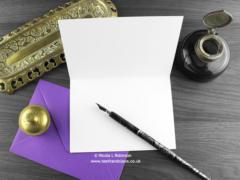 Crow greeting card, shown open and flat with blank inside, on a purple envelope, alongside vintage Victorian pen and writing desk accessories. Design and illustration © Nicola L Robinson | Teeth and Claws