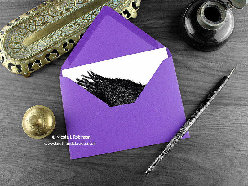 Crow greeting card, shown lying flat, with my original pen and ink illustration of a crow, inside an open purple envelope. With vintage Victorian pen and writing desk accessories. Design and illustration © Nicola L Robinson | Teeth and Claws