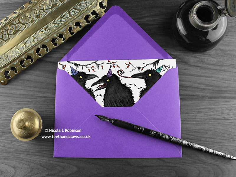 Crows birthday card, shown lying flat, with my original pen and ink illustration of crows, inside an open purple envelope. With vintage Victorian pen and writing desk accessories. Design and illustration © Nicola L Robinson | Teeth and Claws