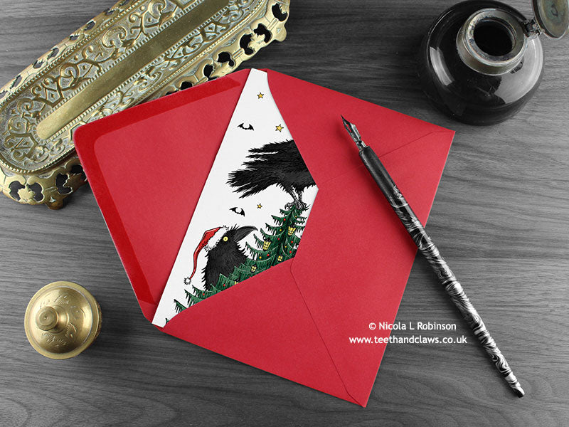 Crow Christmas card, shown lying flat, with my original pen and ink illustration of festive crows, inside an open red envelope. With vintage Victorian pen and writing desk accessories. Design and illustration © Nicola L Robinson | Teeth and Claws